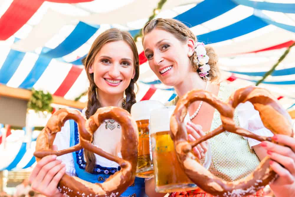 Two adult women dressed in traditional German outfits, holding out large pretzels on a sunny day, having fun.