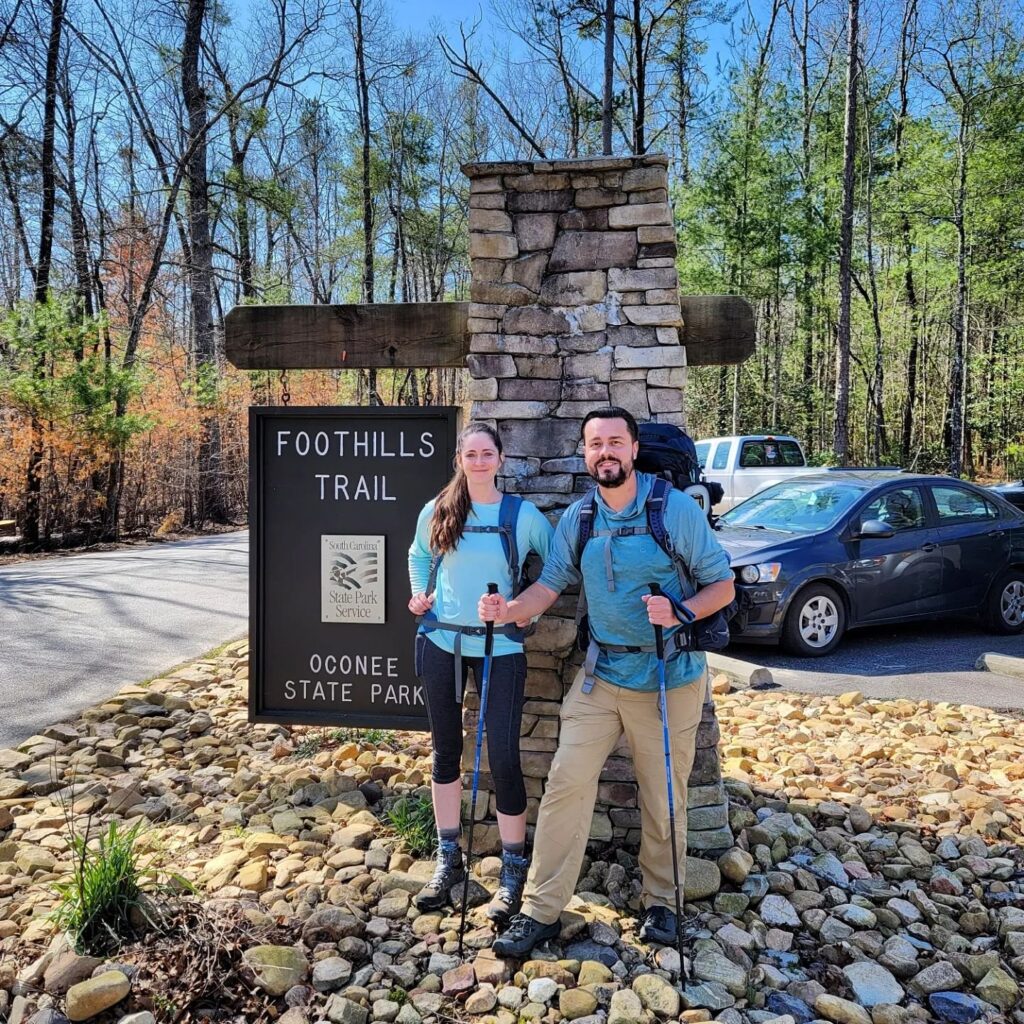 Man and woman in hiking gear, standing in front of Foothills Trail Oconee State Park sign.